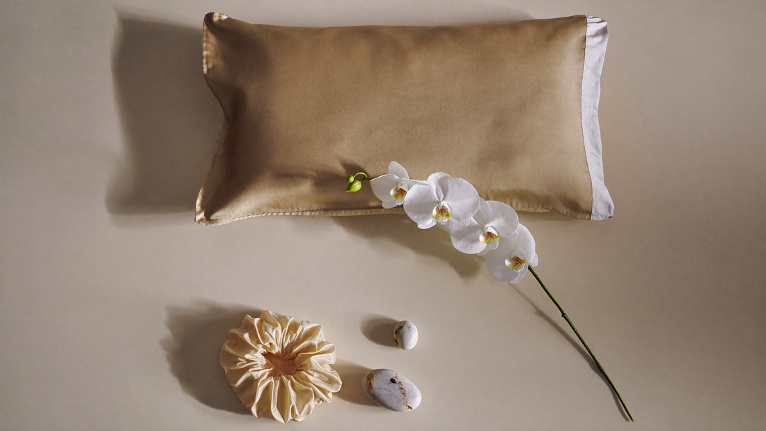 Flat lay de taie d'oreiller et chouchou en soie premium sur lin crème, avec fleurs blanches et pierres lisses dans une lumière naturelle douce