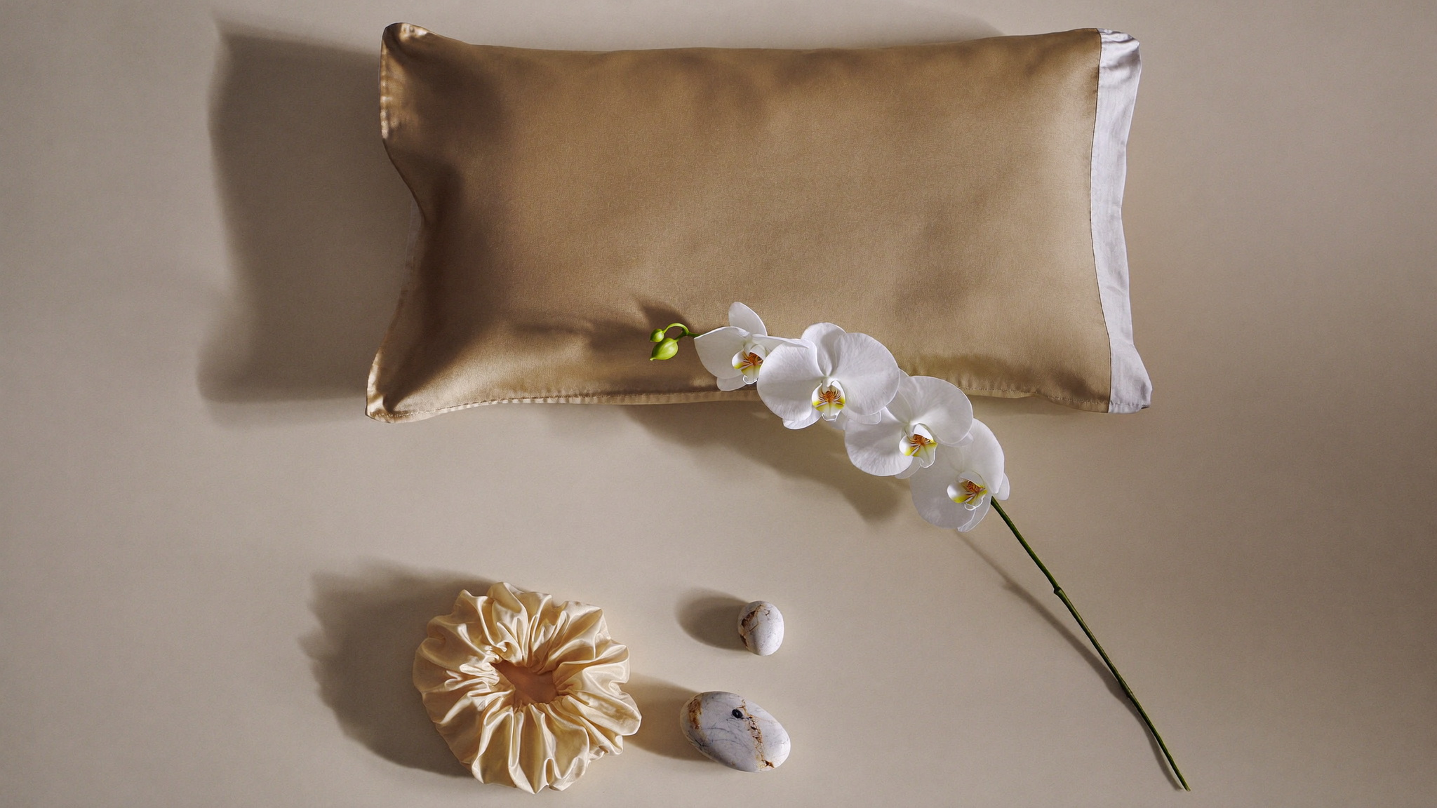 Flat lay de taie d'oreiller et chouchou en soie premium sur lin crème, avec fleurs blanches et pierres lisses dans une lumière naturelle douce