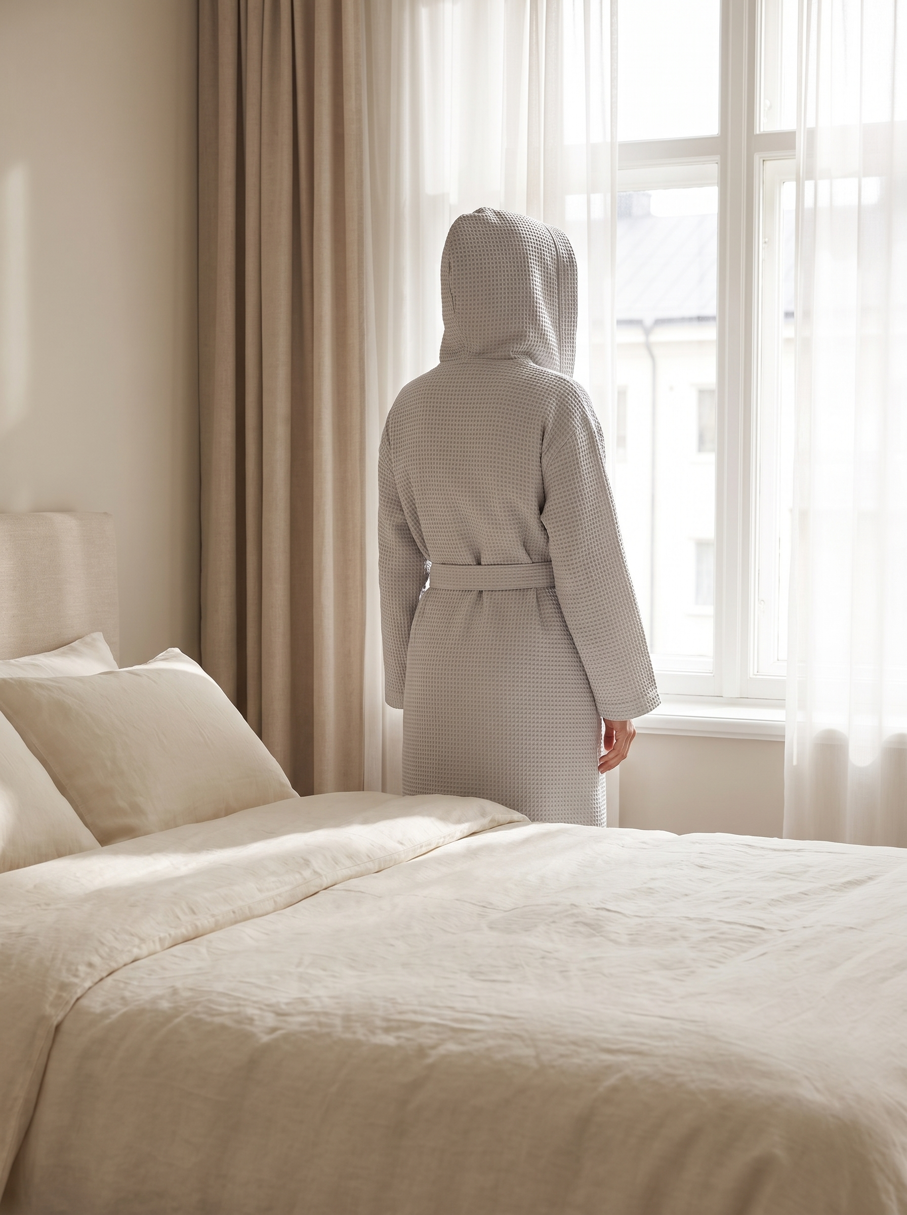 Peignoir en coton gaufré gris clair porté capuche relevée, silhouette de dos face à la fenêtre